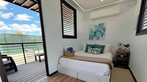 Slaapkamer met een bed, een nachtkastje, een wandairconditioner, een raam met zonwering en uitzicht op een tennisbaan.
