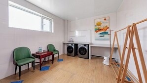 A laundry room with two green chairs, a table with a water bottle, two washing machines, a drying rack, and a painting on the wall.