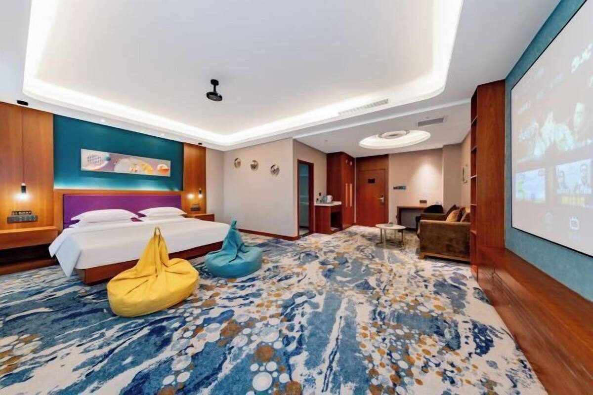 Une chambre d’hôtel moderne dotée d’un grand lit, d’un coin salon avec un canapé et des fauteuils, d’une petite table et d’un grand écran diffusant du contenu.