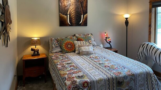 Un dormitorio con cama, ventilador de techo, un cuadro de elefante en la pared, una mesita de noche con lámpara y una lámpara de pie.