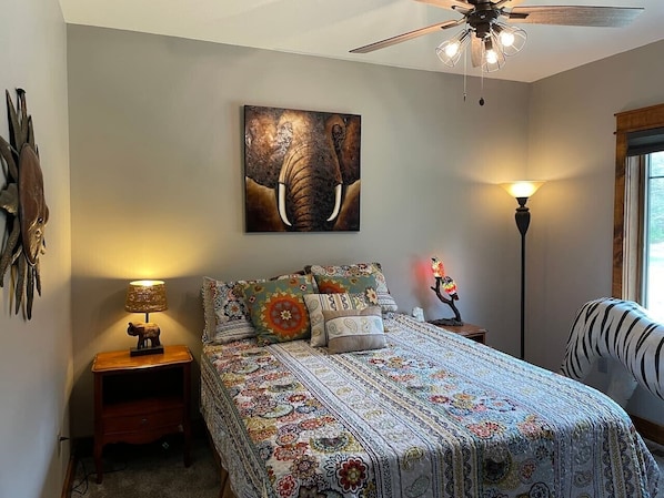 A bedroom with a bed, a ceiling fan, a wall-mounted elephant painting, a nightstand with a lamp, and a floor lamp.