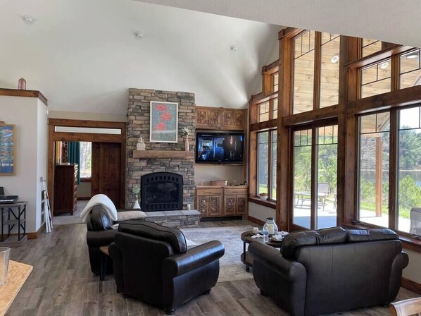 A spacious living room with a stone fireplace, leather sofas, a flat-screen TV, and large windows offering a view of the outdoors. - Woodsy and Private- Dream House on a 7 Acre Pond (Paradise)