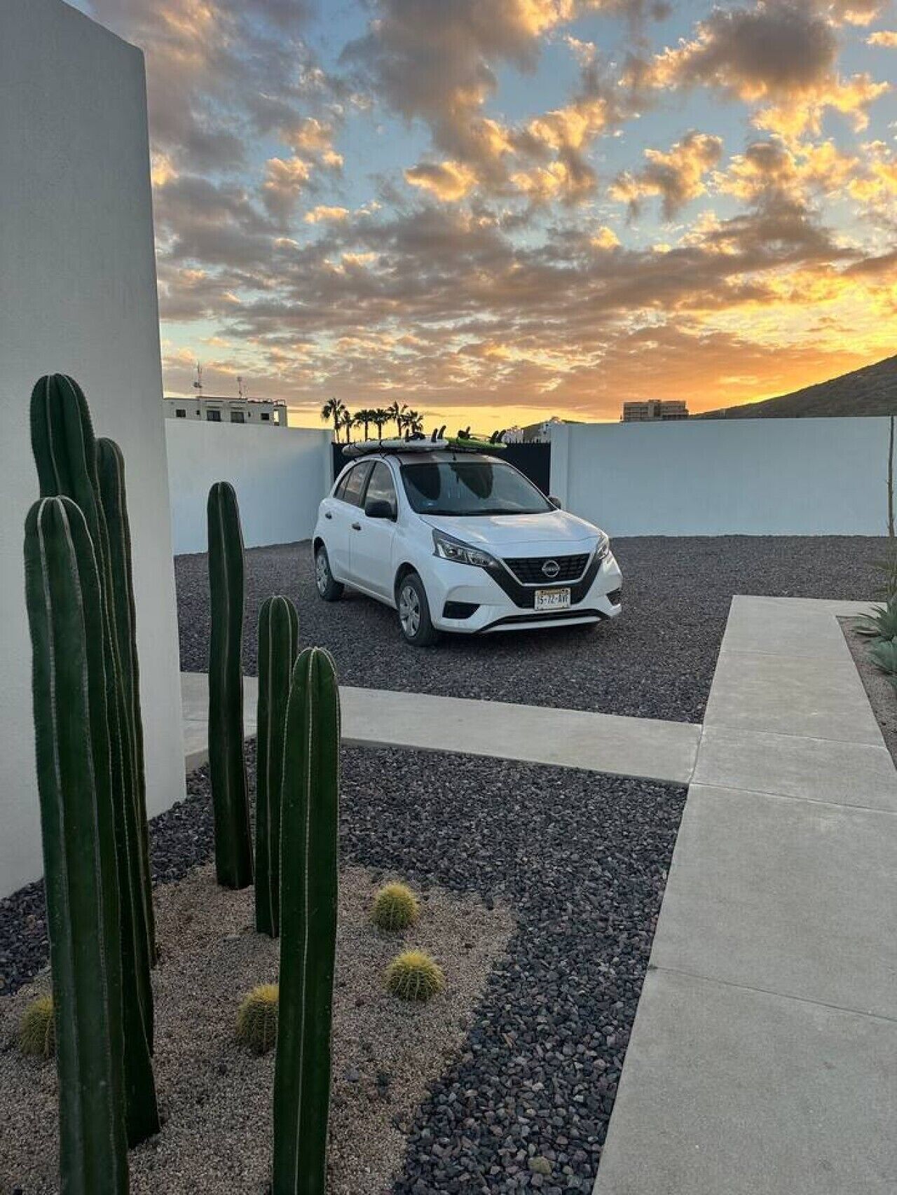 A white car parked on a gravel driveway with cacti in the foreground and a sunset in the background.