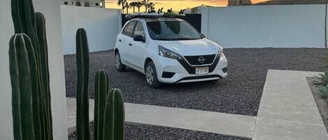A white car parked on a gravel driveway with cacti in the foreground and a sunset in the background.