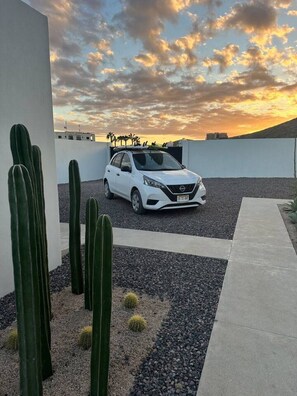 Un coche blanco estacionado en un camino de grava, con cactus en primer plano y una puesta de sol al fondo.