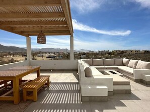 Eine Terrasse im Freien mit einem Holztisch und -bänken, einer gepolsterten Sitzgruppe und einer hängenden Ampel.