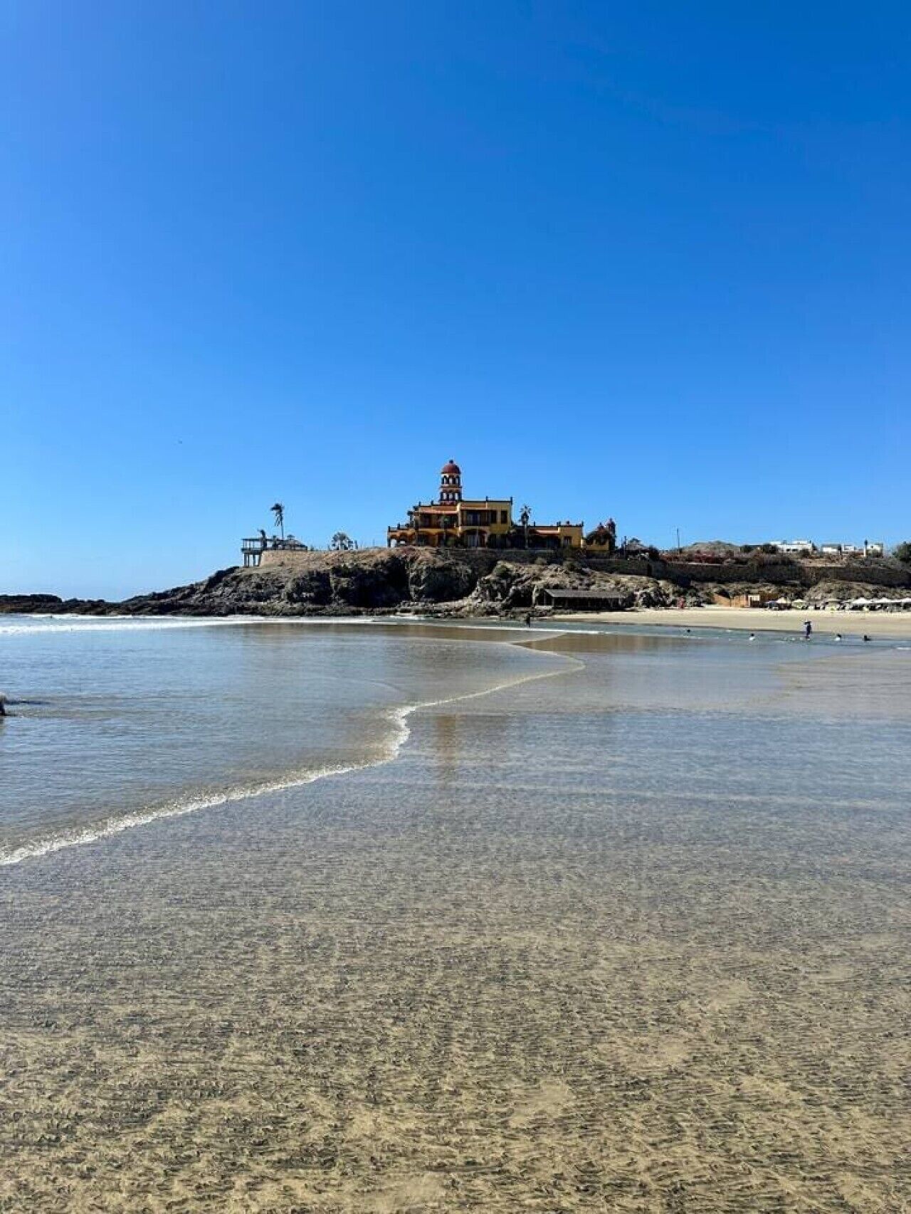 A coastal building with a dome on top, situated on a rocky outcrop near a sandy beach.