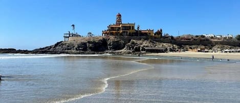 A coastal building with a dome on top, situated on a rocky outcrop near a sandy beach.