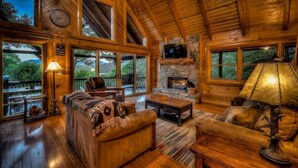 A cozy cabin interior with a stone fireplace, wooden furniture, and large windows offering a view of the outdoors. - Bearway to Heaven Lake View Fire Pit Game Room (Blue Ridge)