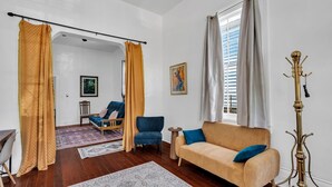 A living room with a sofa, armchair, and a small table. There is a window with curtains and a coat rack.