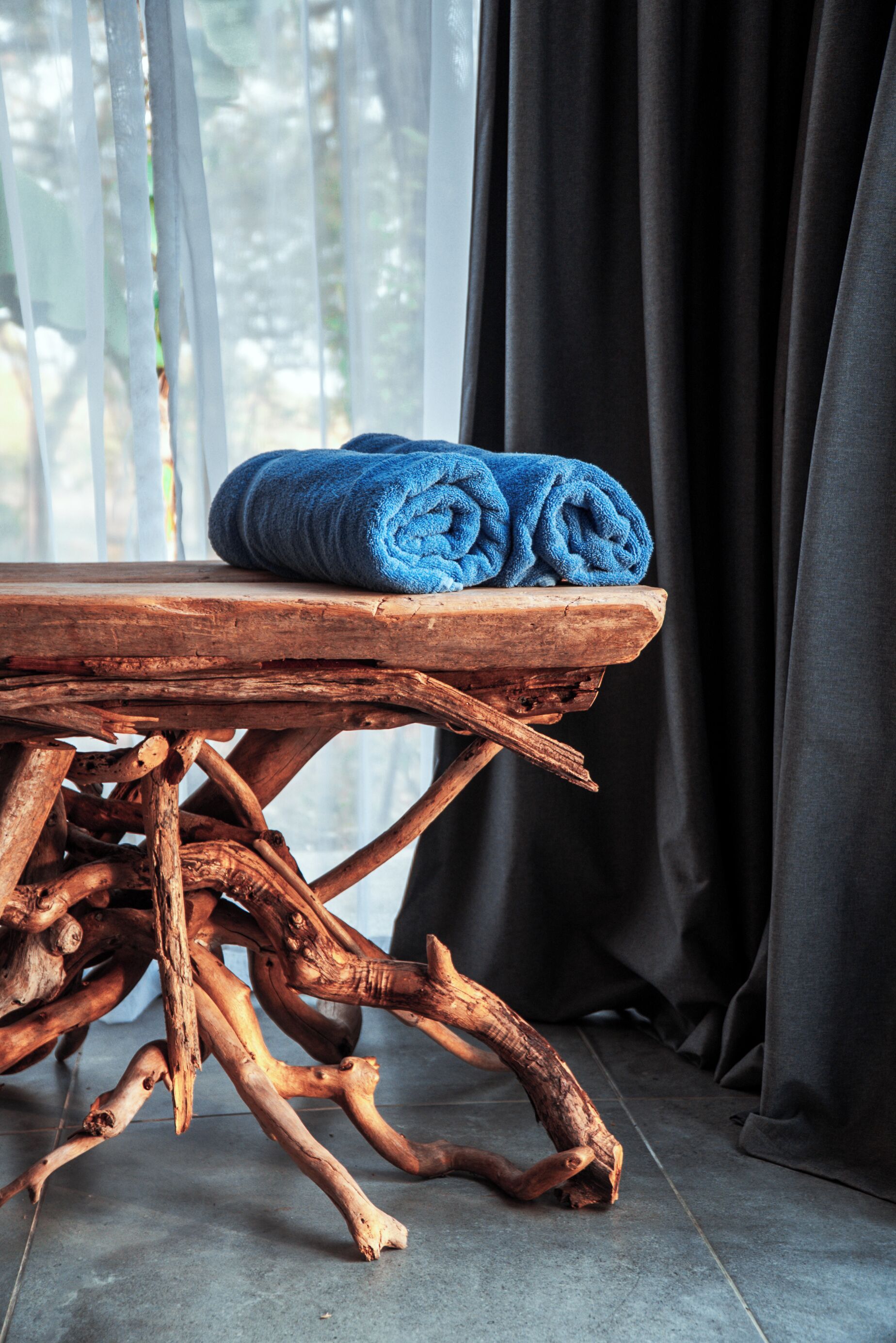 Une table en bois sur laquelle est posée une serviette bleue roulée, dans une pièce dotée d’une fenêtre et de rideaux.
