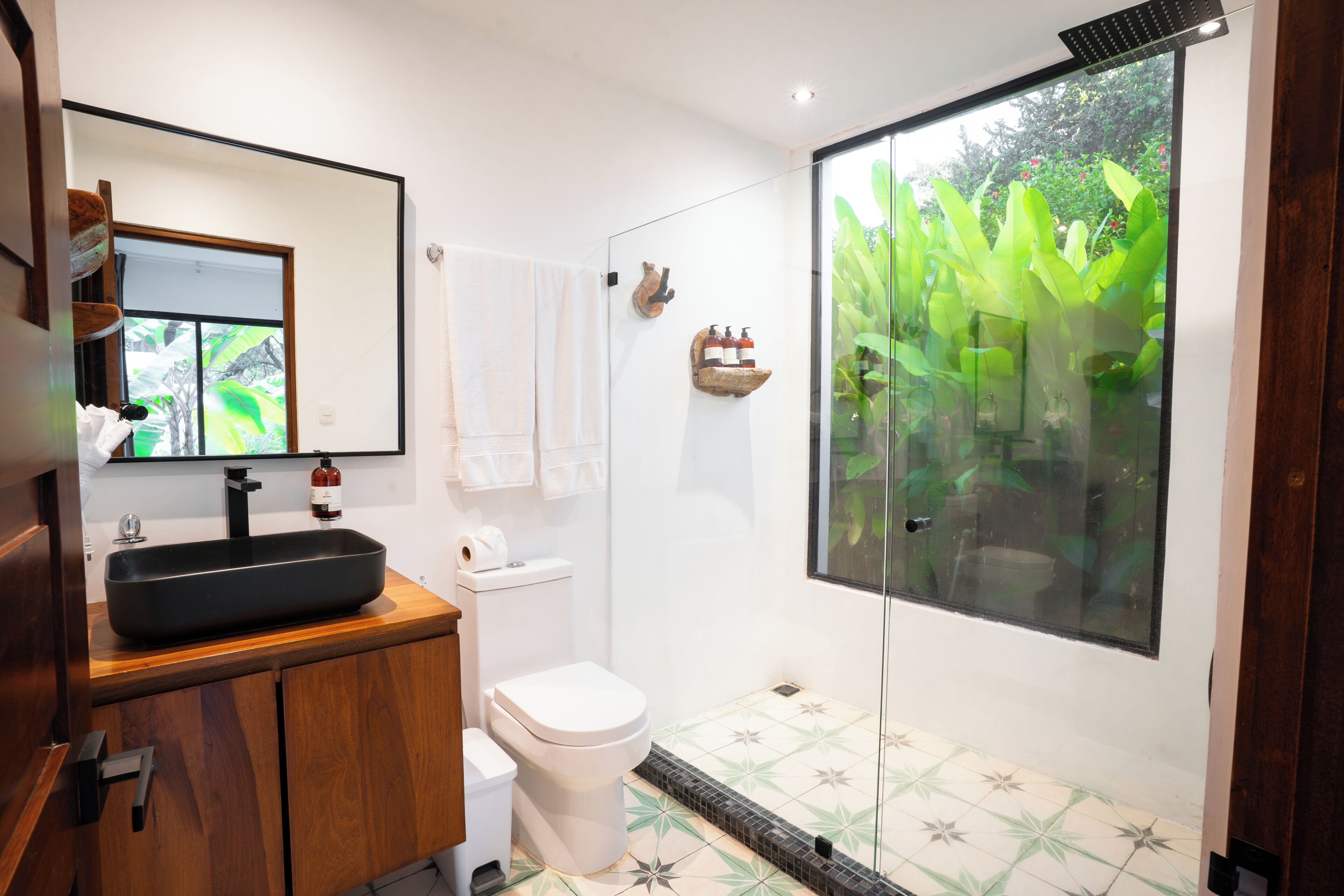 Un baño moderno con un lavabo negro, inodoro blanco y cabina de ducha de vidrio con vistas a zonas verdes.