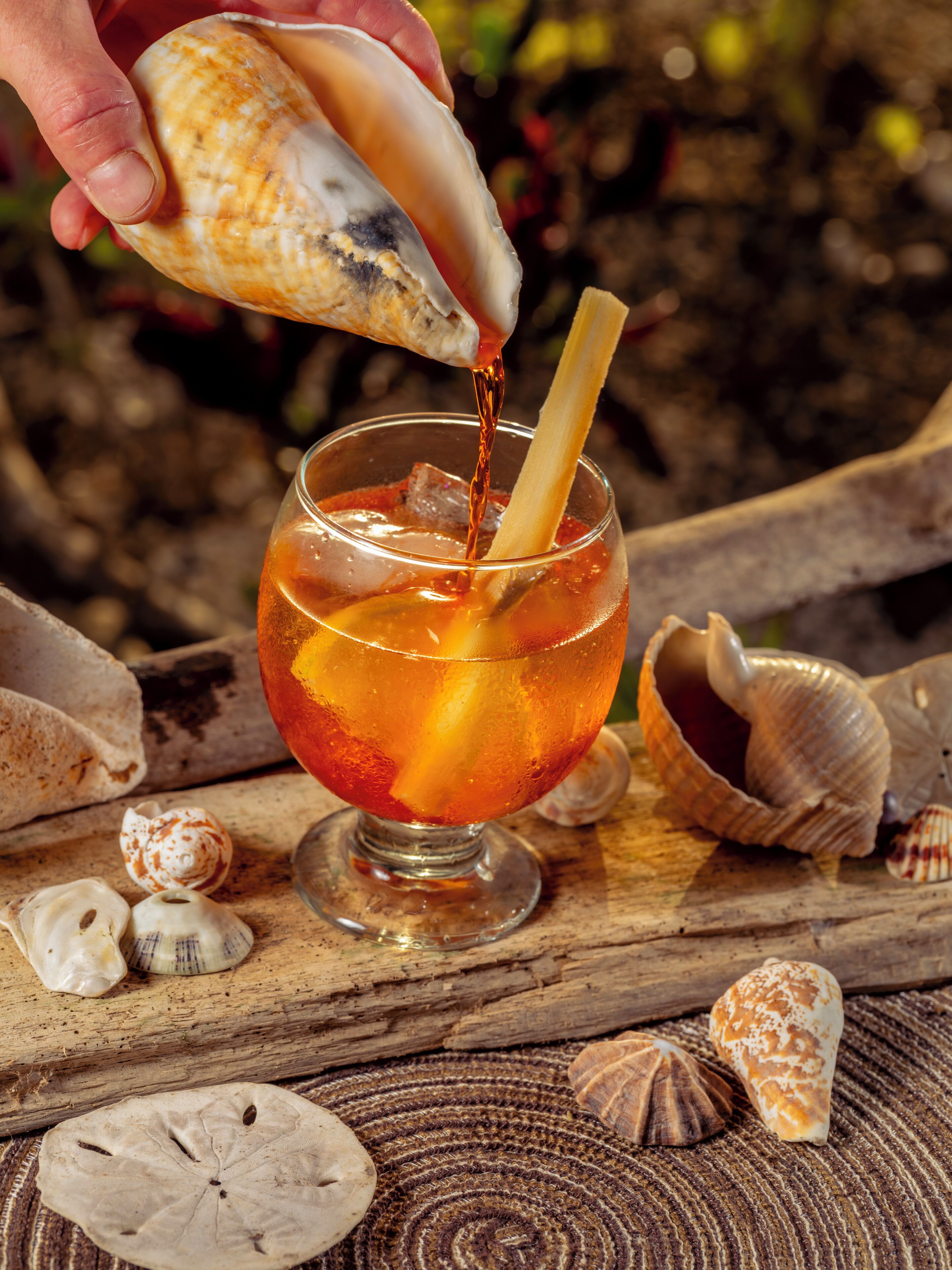 A glass of amber liquid with a straw, surrounded by seashells on a wooden surface.