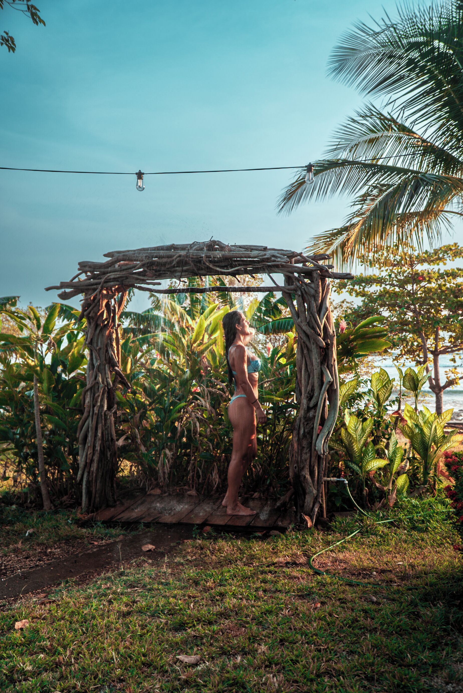 Une personne se tient sous une structure en bois, dans un cadre tropical caractérisé par une végétation luxuriante et un ciel dégagé.