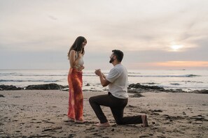 Escena en una playa con una persona arrodillada proponiendo matrimonio a otra de pie.