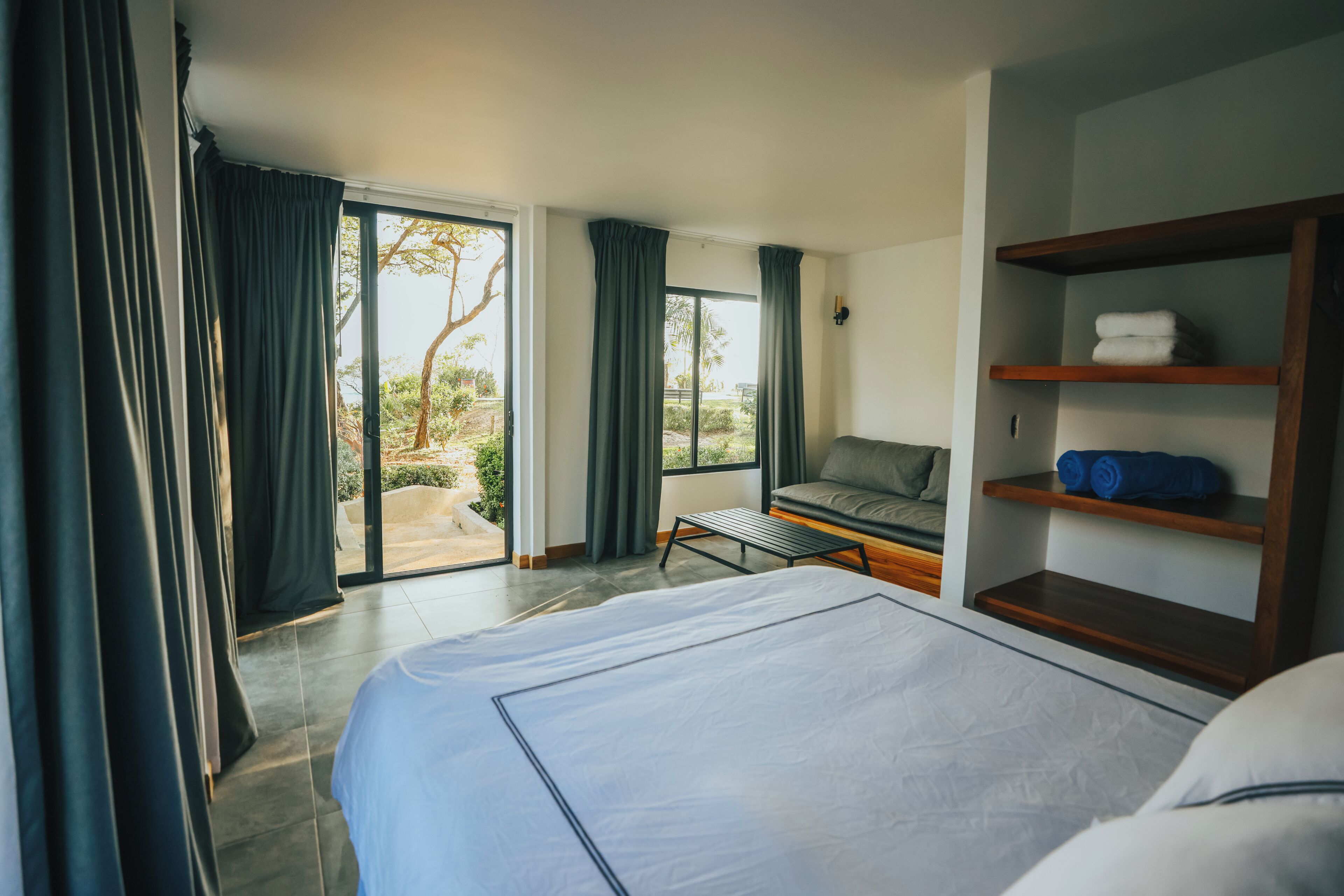A hotel room with a bed, a seating area with a view, and a shelf with towels.