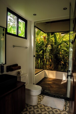 Un baño con inodoro, lavamanos y cabina de ducha de vidrio con vistas a follaje tropical.