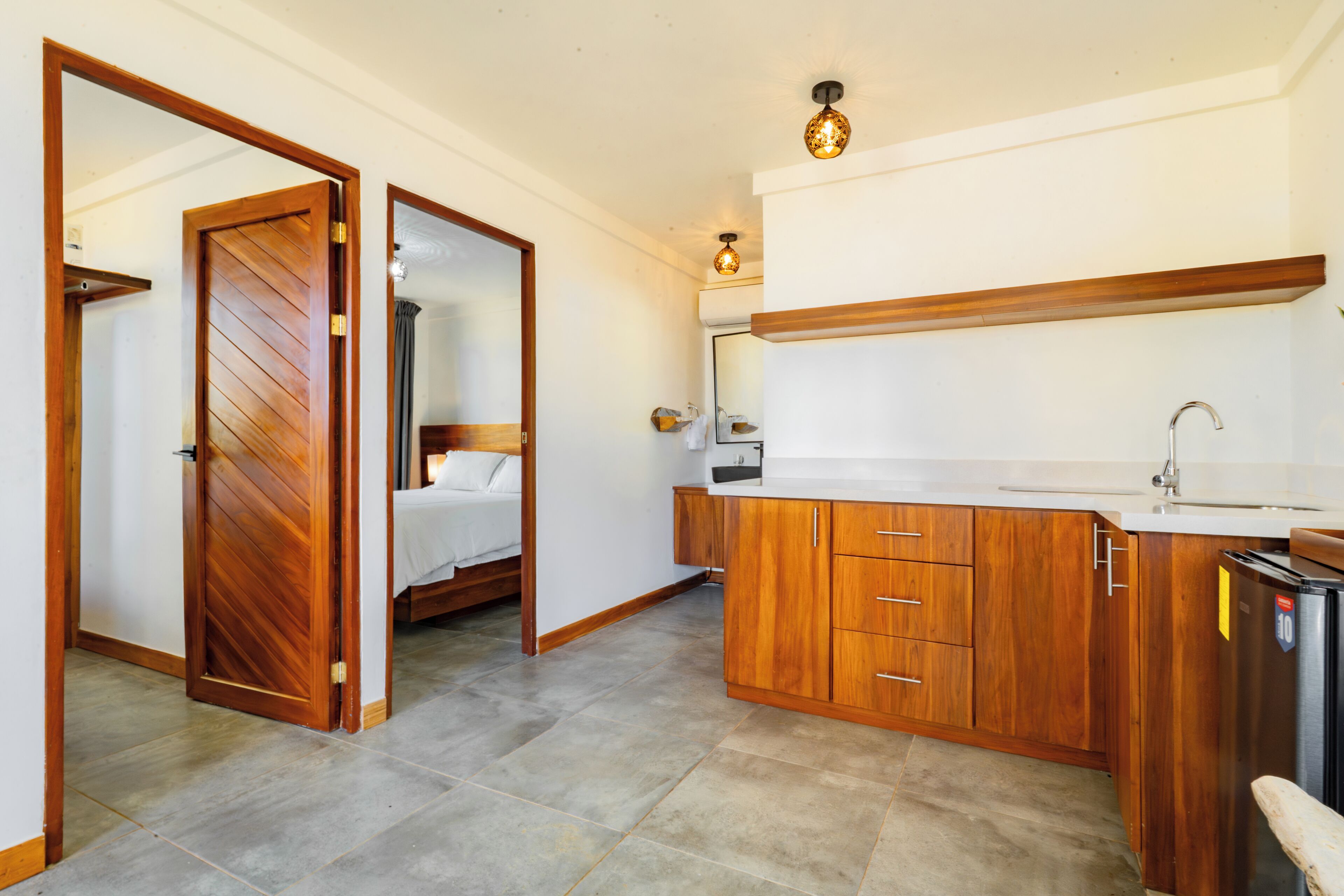 A compact hotel room with a wooden wardrobe, a bed, a small bathroom with a mirror, and a kitchenette with a sink and a refrigerator.