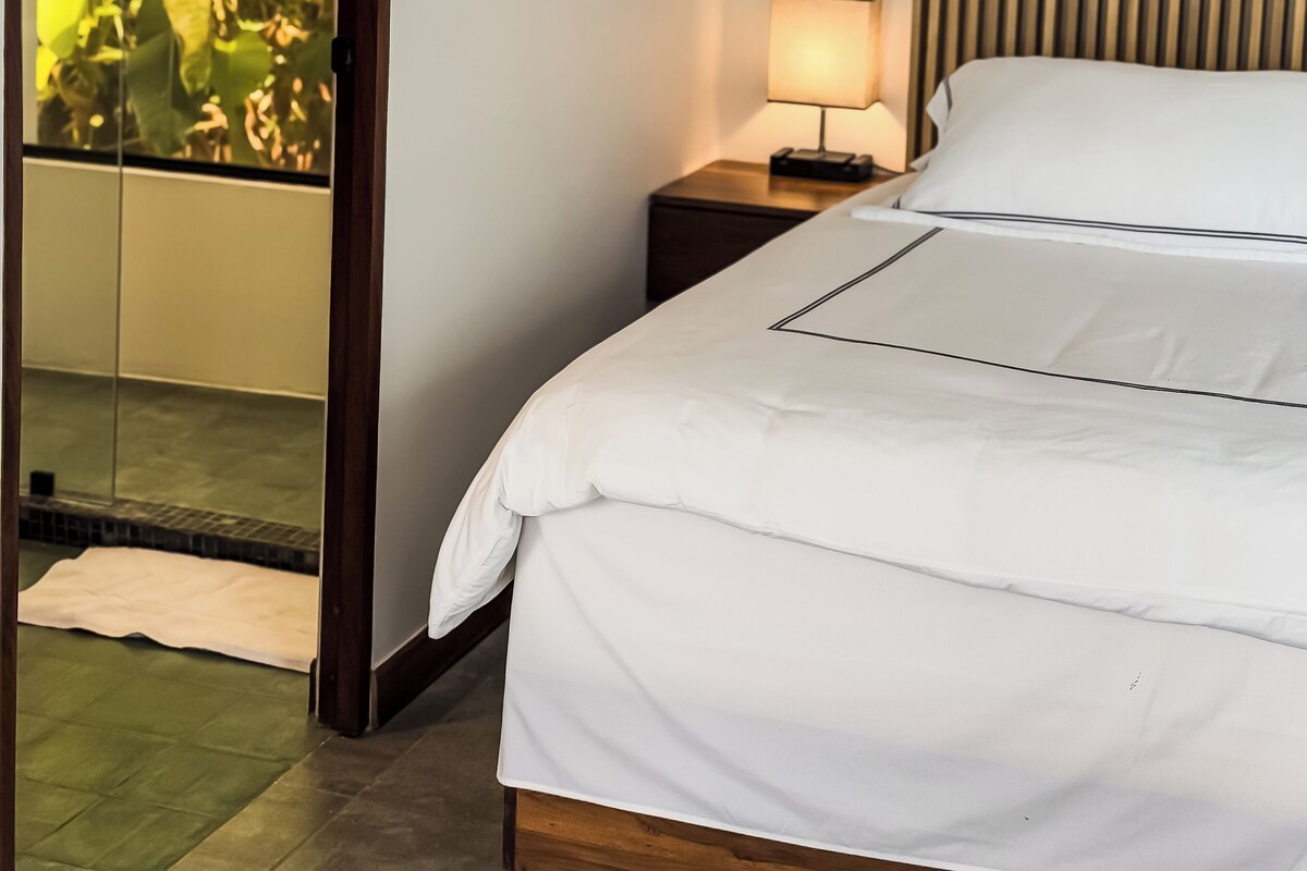 Habitación de hotel con cama, mesita de noche con lámpara, unidad de aire acondicionado y vista a zonas verdes a través de una puerta de cristal.
