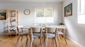 A dining area with a round table, four chairs, a wooden bookshelf, a wall clock, and a window with blinds.