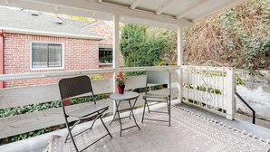 A covered patio area with two metal chairs and a small round table, a brick building in the background, and a white fence with climbing plants.