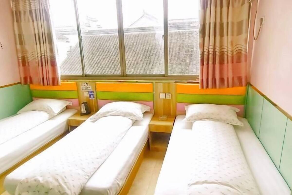 A room with two single beds, a window with curtains, a small table with a lamp, and a wall-mounted phone.