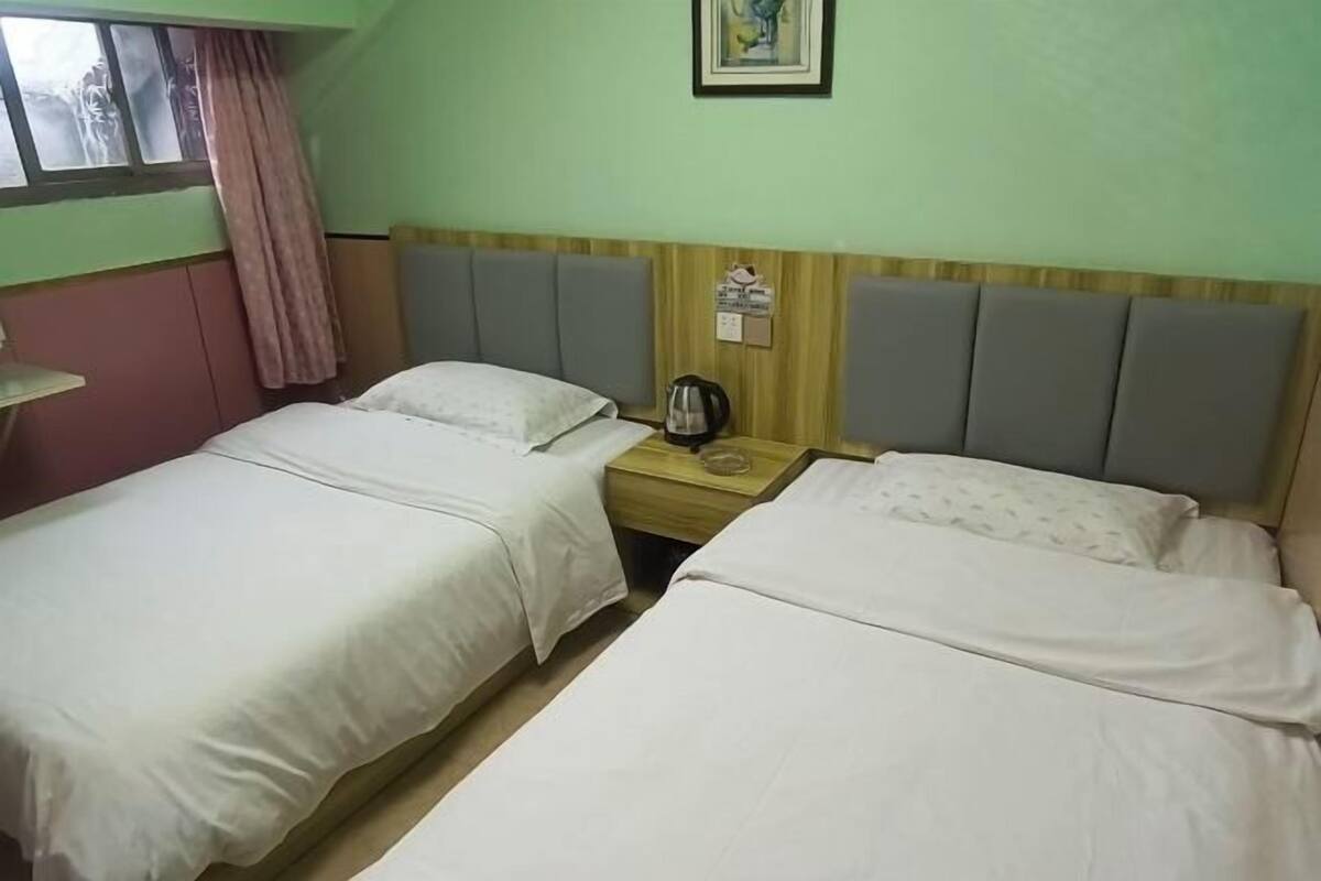 A hotel room with two single beds, a wooden nightstand with a kettle, and a window with curtains.