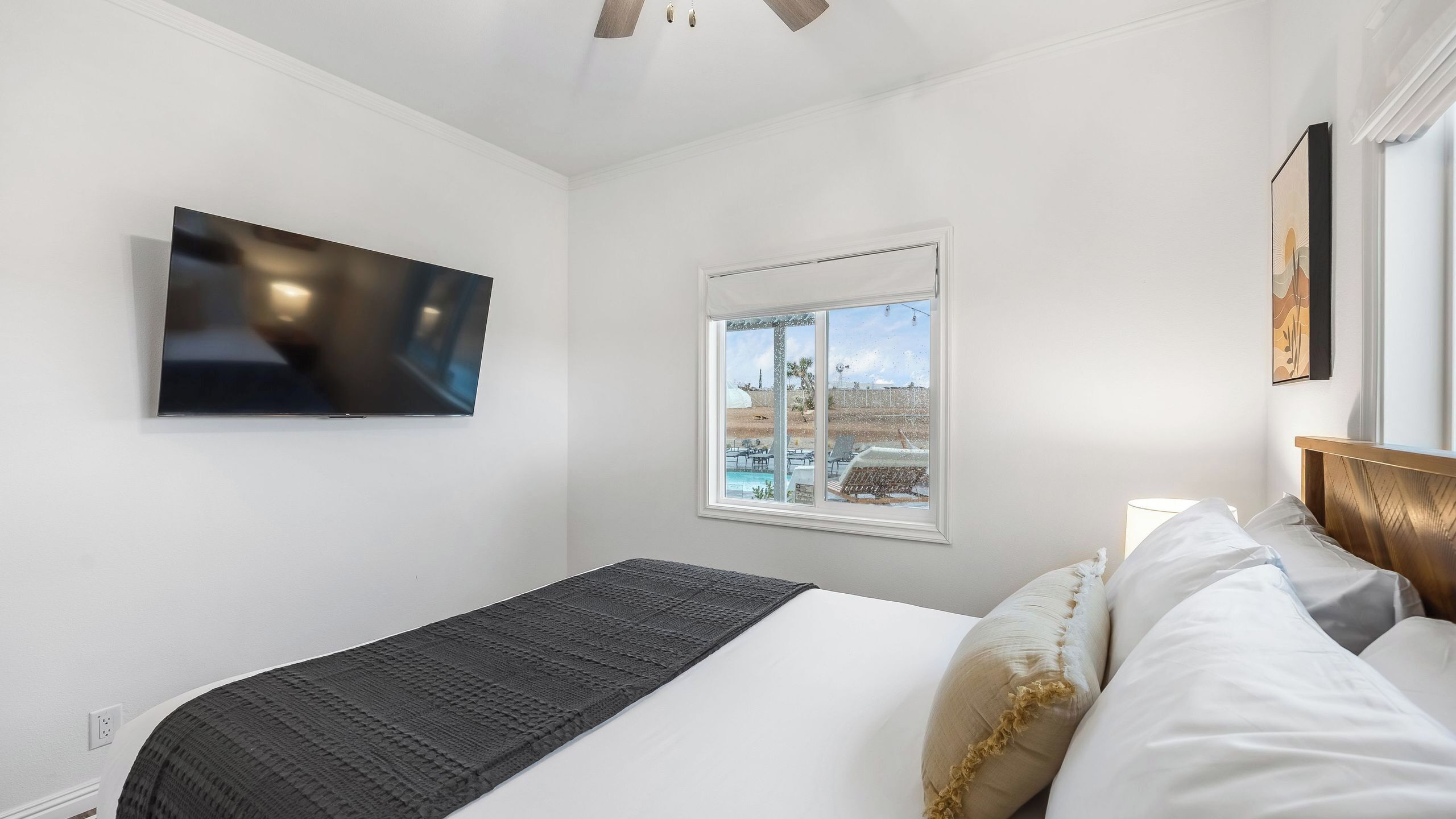 A bedroom with a bed, a TV mounted on the wall, a window with a view of a pool and palm trees, and a ceiling fan.