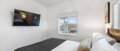 A bedroom with a bed, a TV mounted on the wall, a window with a view of a pool and palm trees, and a ceiling fan.