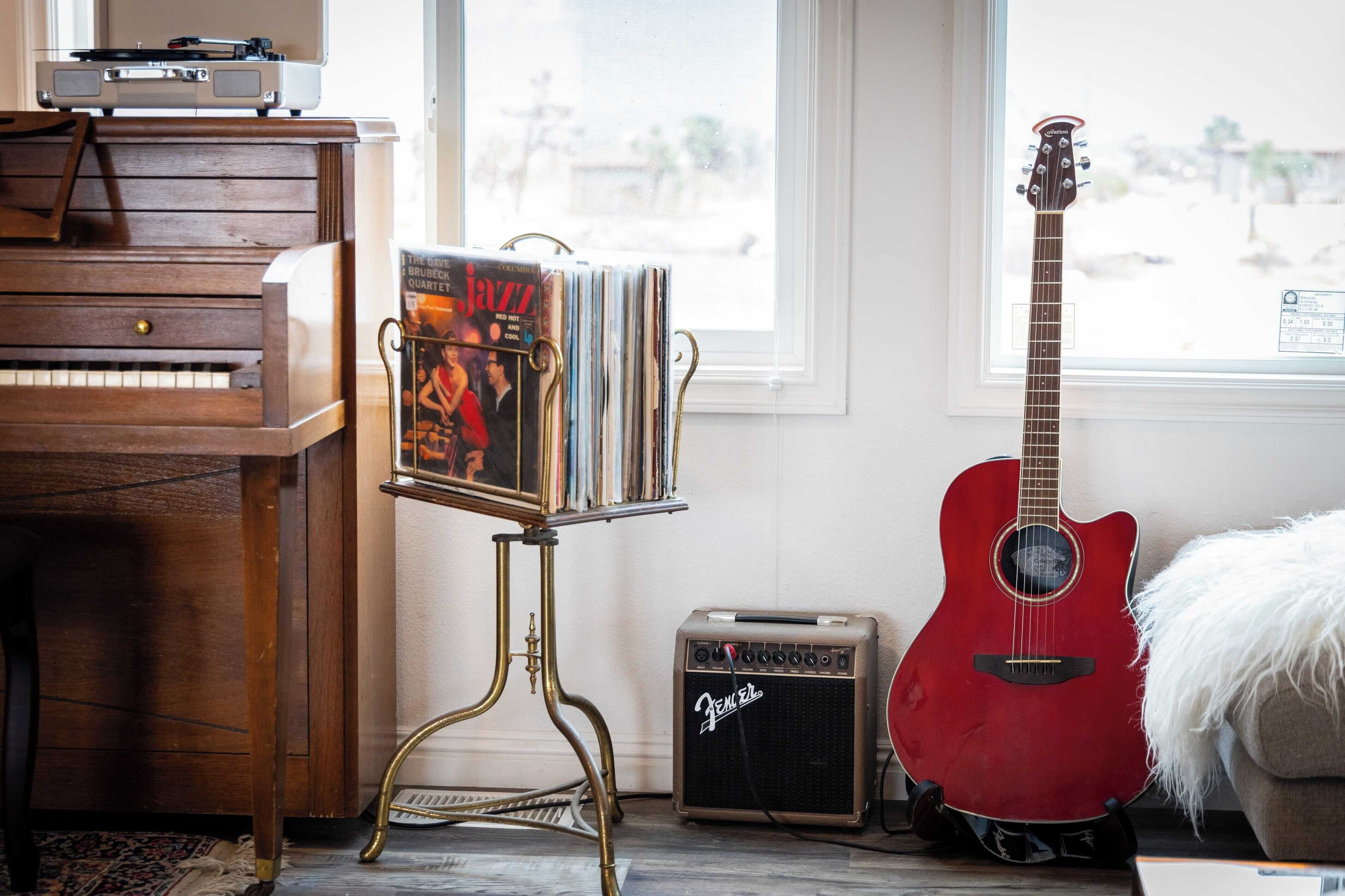 A room with a wooden piano, a Fender amplifier, a red guitar, and a rack of vinyl records.