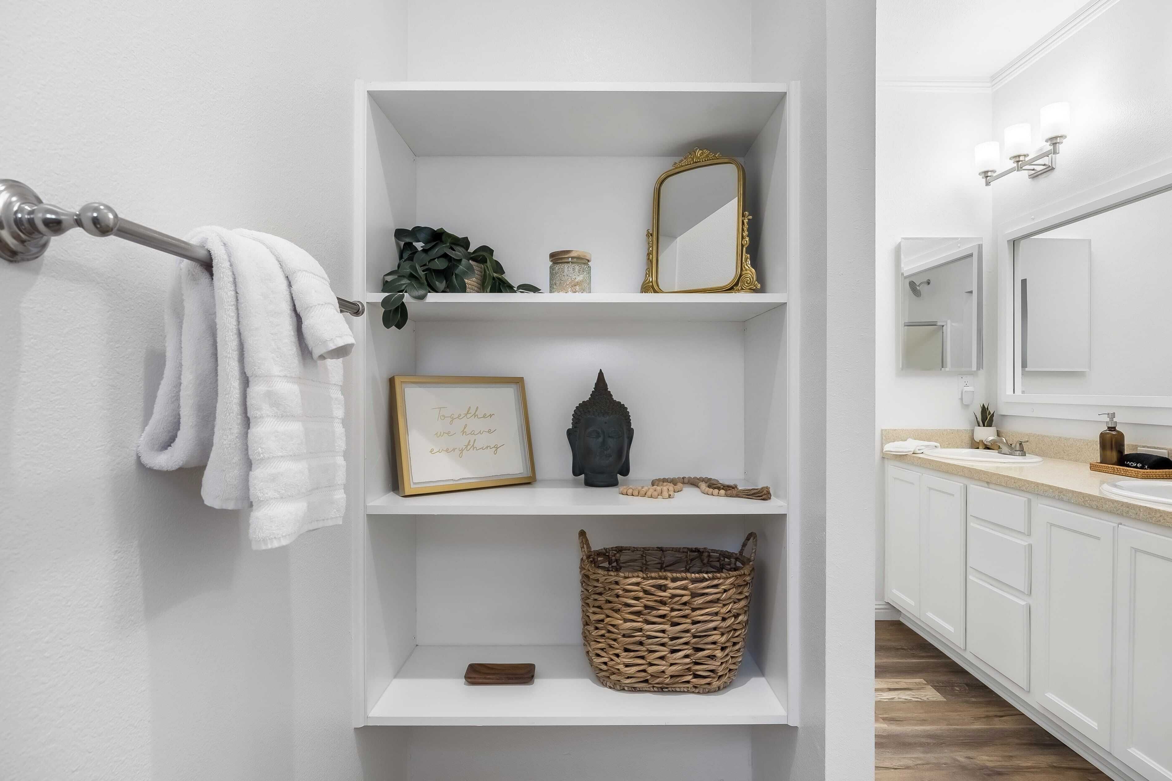 A bathroom with white shelves, a towel rack, a framed quote, a Buddha statue, a woven basket, and a mirror.
