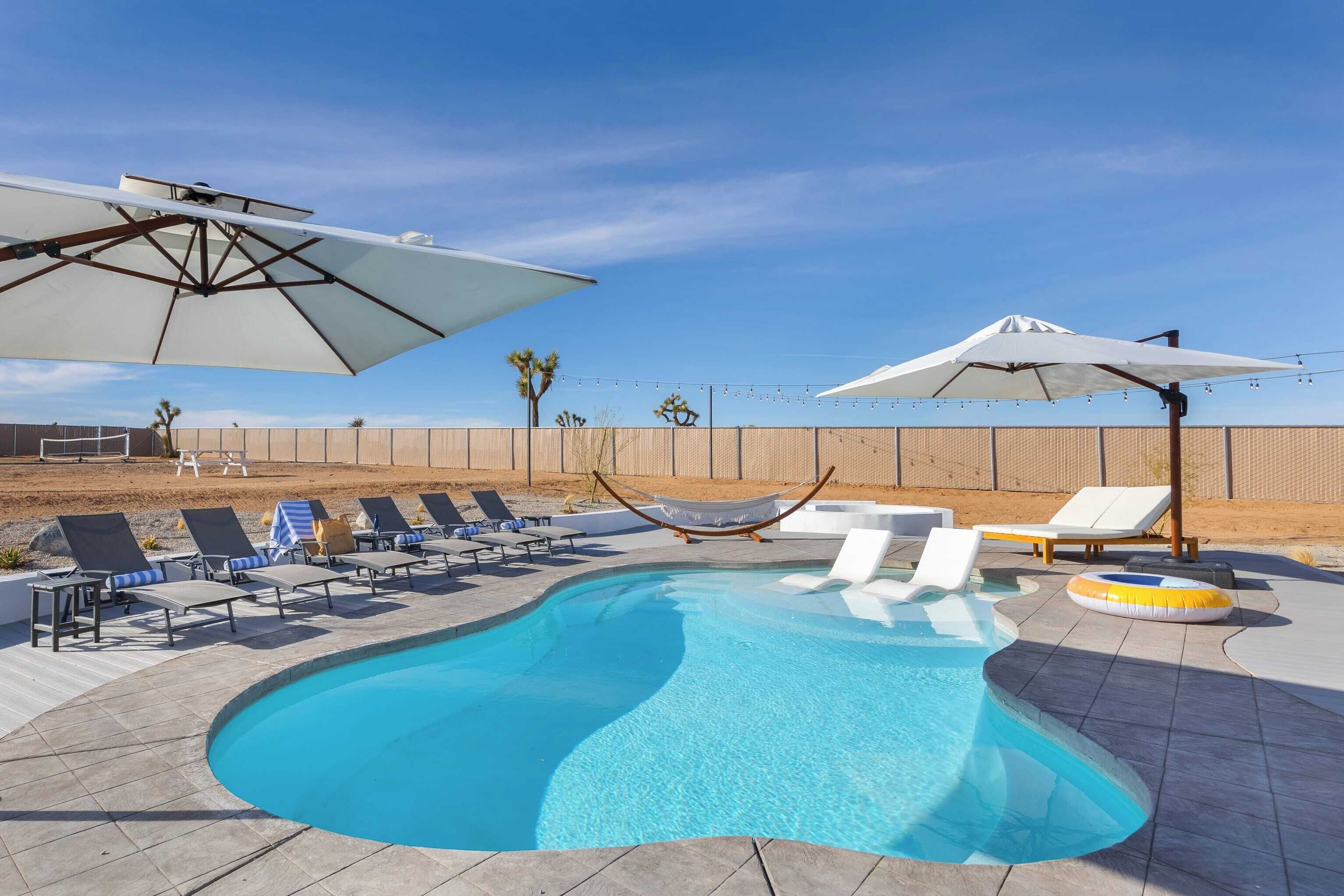 A swimming pool with lounge chairs and umbrellas, surrounded by a fence and desert landscape.