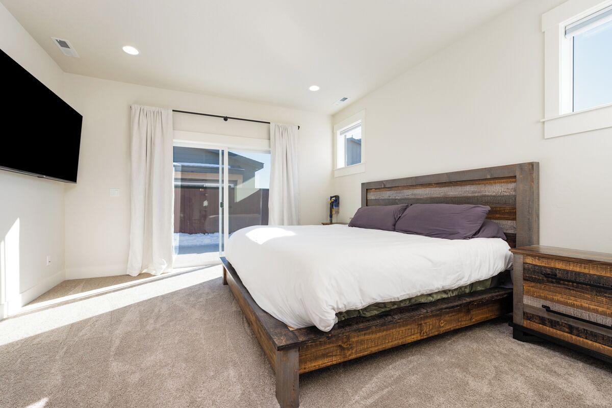 A bedroom with a large bed, a wooden nightstand, a flat-screen TV mounted on the wall, and a sliding glass door leading to an outdoor area.