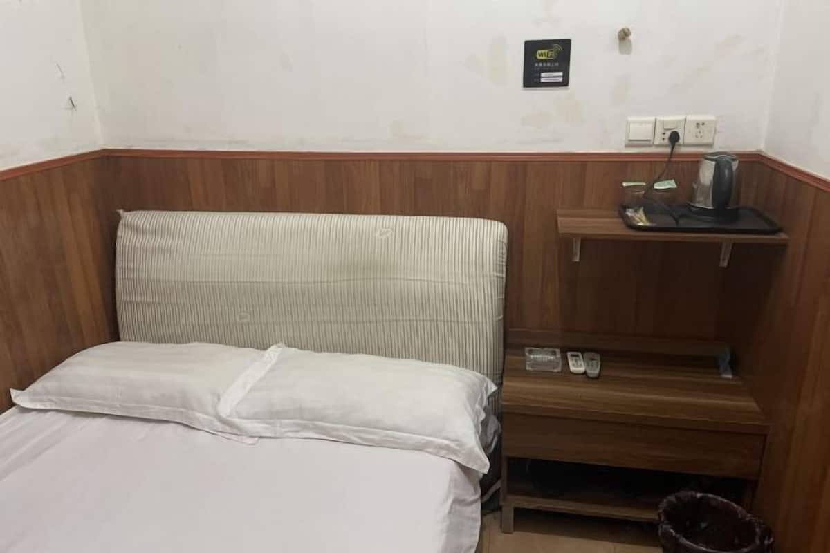 Una habitación de hotel con cama, cabecera de madera, mesita de noche con tetera y un cubo de basura.