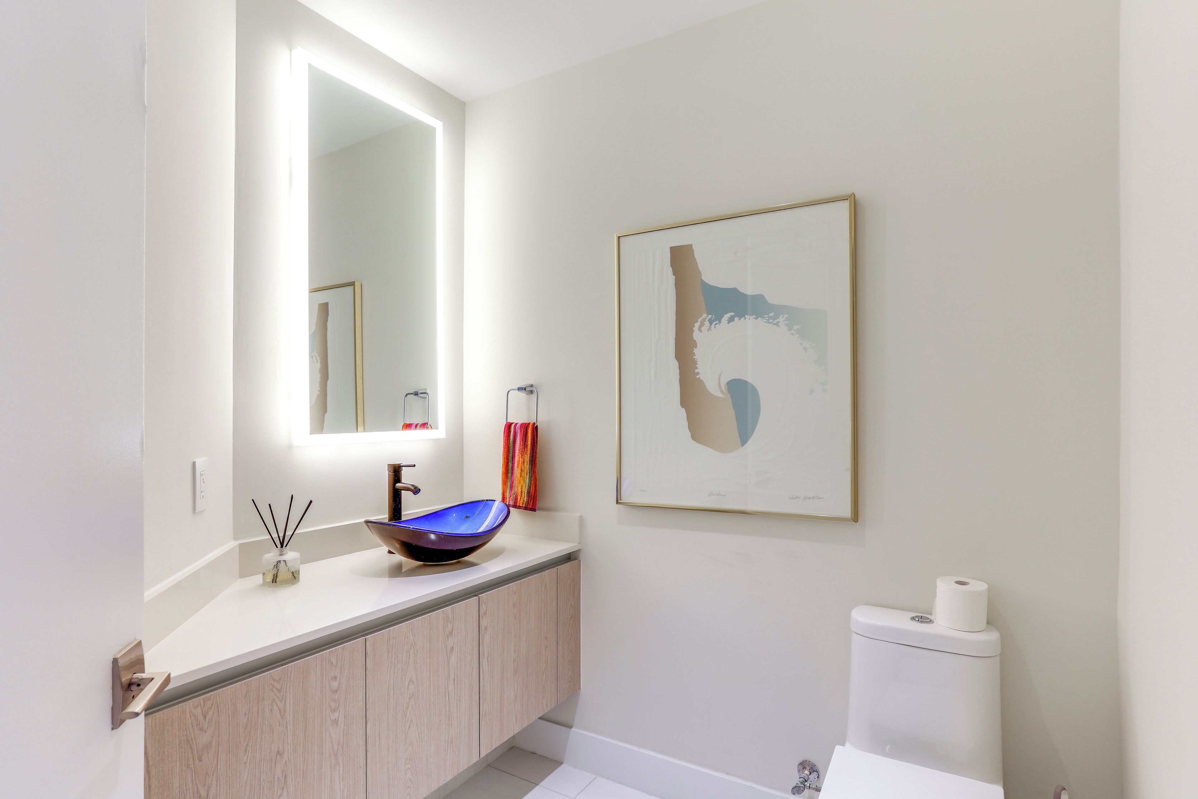 Une salle de bain moderne avec un lavabo blanc, un miroir, des toilettes et une œuvre d’art encadrée accrochée au mur.