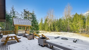 Una terraza de madera con una mesa de picnic y bancos, rodeada de suelo y árboles cubiertos de nieve.