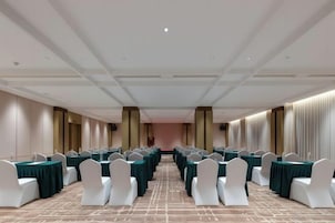 Un salón de conferencias con filas de mesas y sillas blancas, una alfombra estampada y un techo alto con iluminación empotrada.
