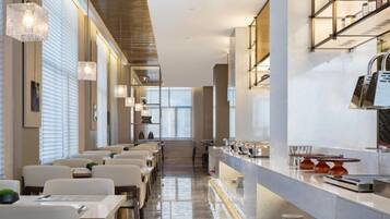 A modern hotel dining area with long tables and chairs, a kitchen counter, and a view of the dining area.