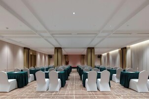 A conference room with rows of tables and chairs, a carpeted floor, and a high ceiling with recessed lighting.