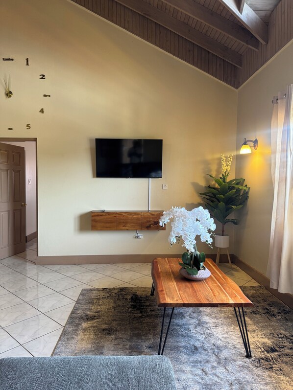 A modern living room with a flat-screen TV mounted on the wall, a wooden bench below it, a potted orchid on a wooden coffee table, and a tiled floor. - 2BR Hip Strip•Ocean View Balcony•Steps to Beach (Montego Bay)