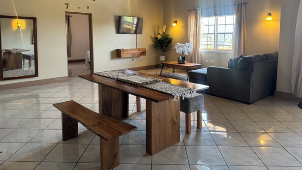 A spacious living area with a wooden table, bench, and grey sofa. A TV is mounted on the wall, and there is a window with curtains. - 2BR Hip Strip•Ocean View Balcony•Steps to Beach (Montego Bay)
