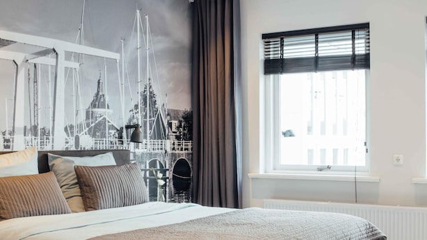 Een slaapkamer met een groot bed, een raam met zonwering, en een muurschildering van een brug en gebouwen aan de wand.