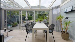 Een oranjerie met glazen dak, een eettafel met stoelen en potplanten.