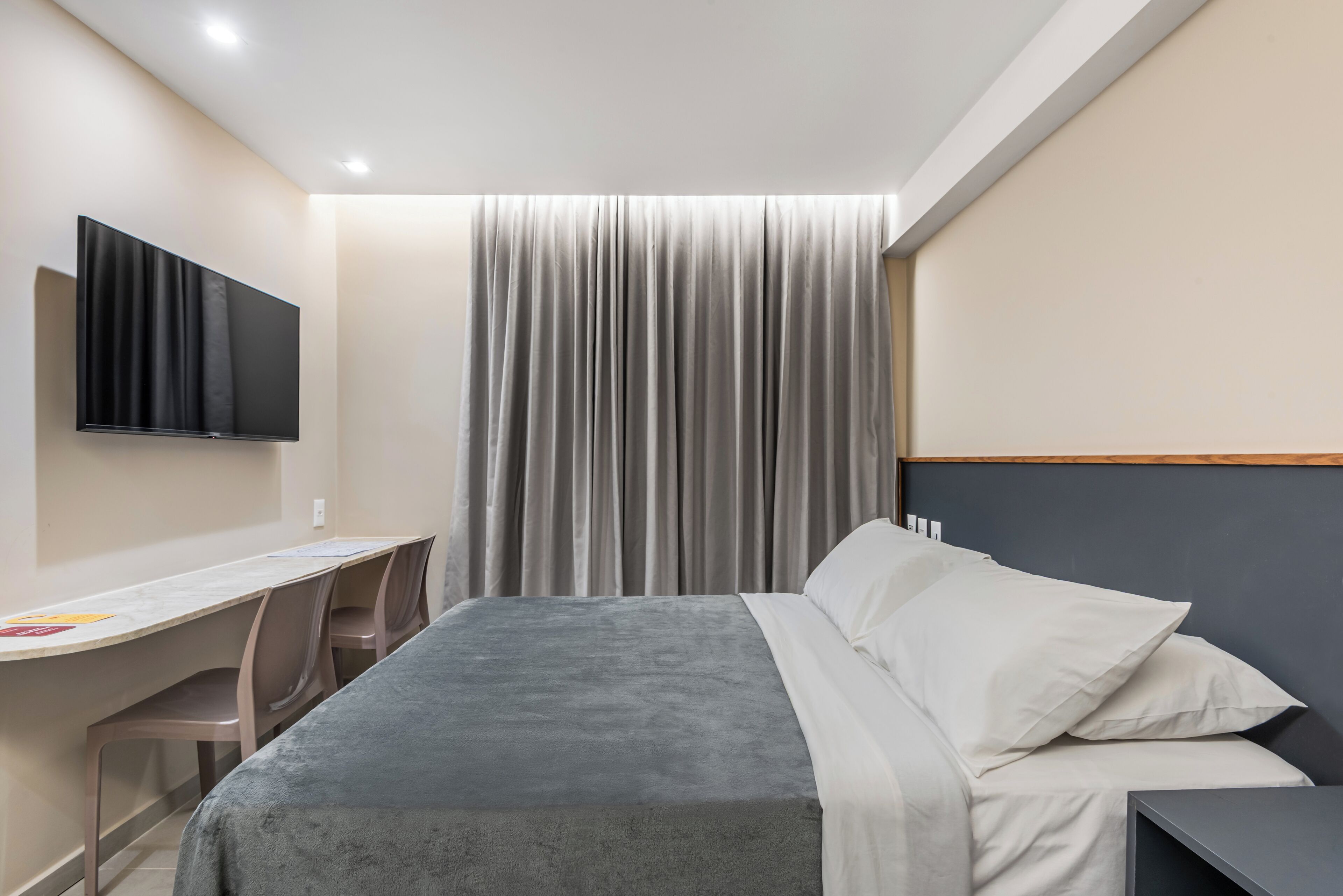 Habitación de hotel con cama, escritorio, silla, televisor montado en la pared y cortina.