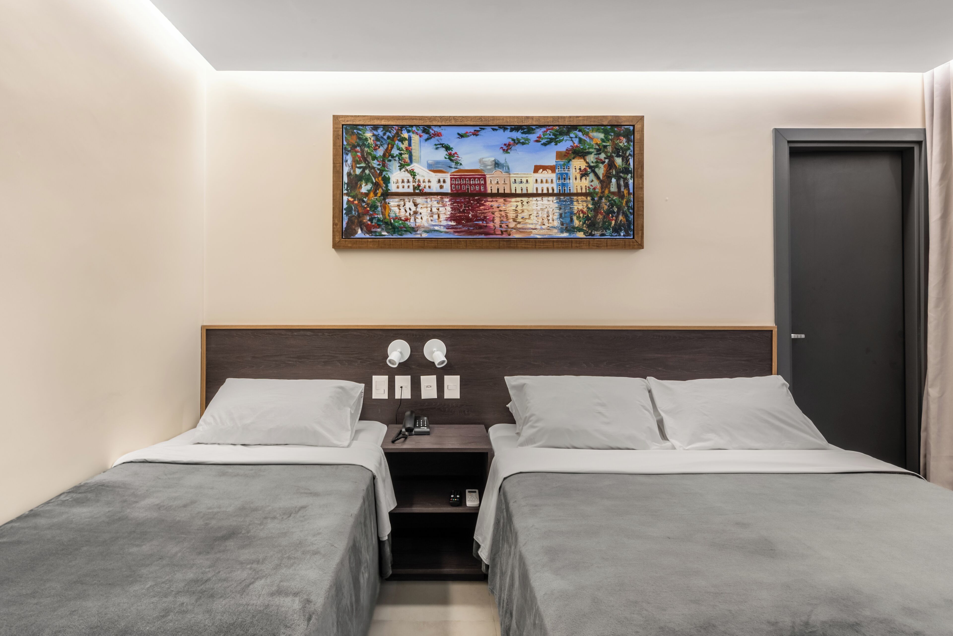 Quarto de hotel com duas camas de solteiro, uma mesa de cabeceira com abajur, um quadro acima da cama e uma porta à direita.