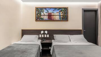 Quarto de hotel com duas camas de solteiro, uma mesa de cabeceira com abajur, um quadro acima da cama e uma porta à direita.