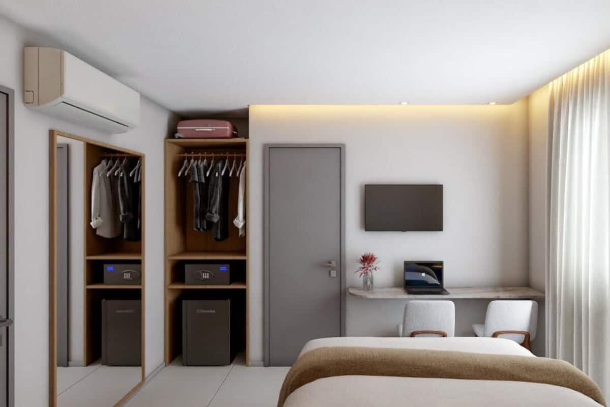 A modern hotel room with a bed, a desk with a laptop, a TV, and a wardrobe with hanging clothes.