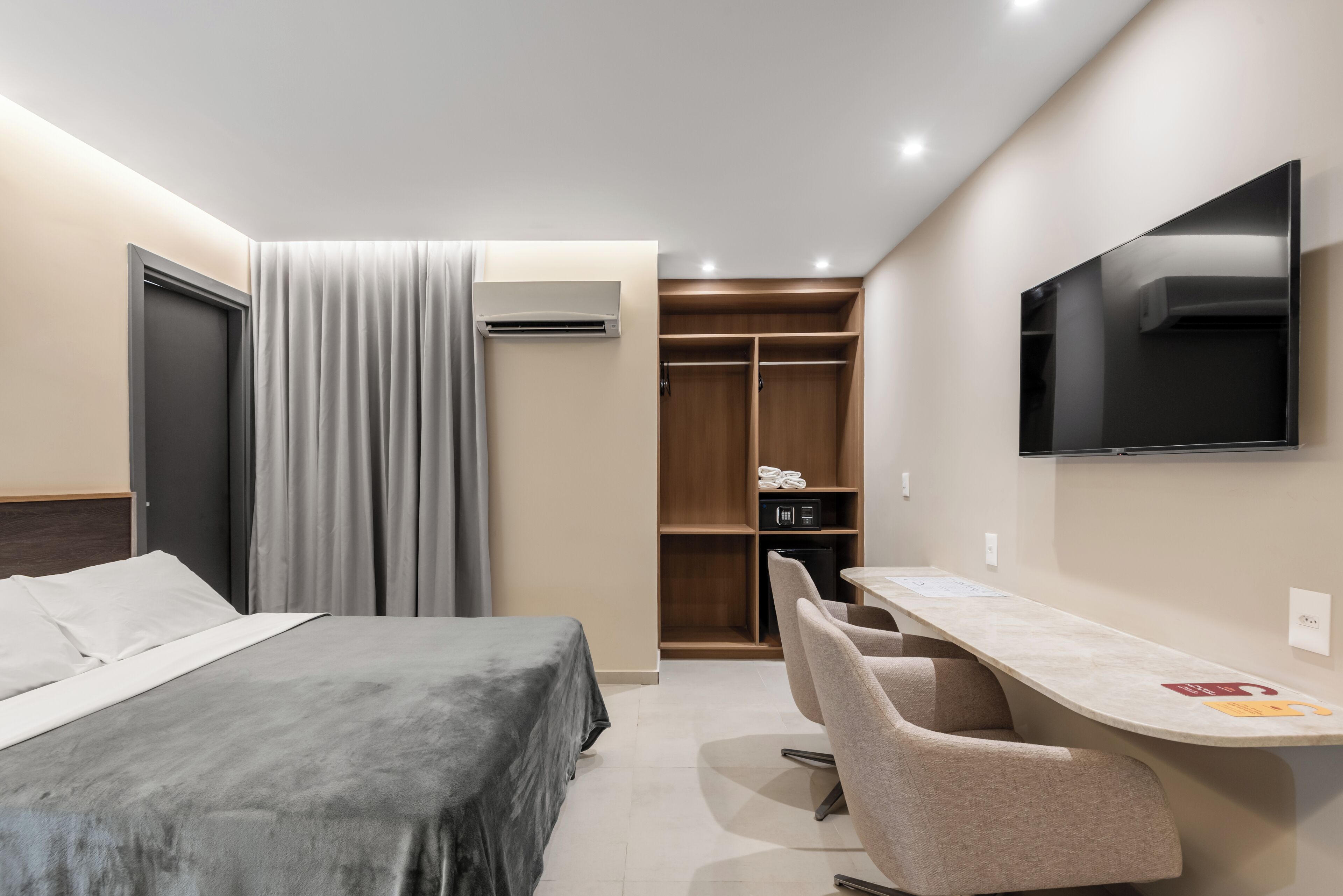 Quarto de hotel com cama, escrivaninha, cadeira, televisão e guarda-roupa.