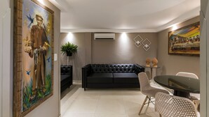 Una sala de estar moderna con un sofá de cuero negro, una mesa redonda de vidrio y dos sillas blancas. Hay un cuadro grande en la pared izquierda y un equipo de aire acondicionado en la pared sobre el sofá.