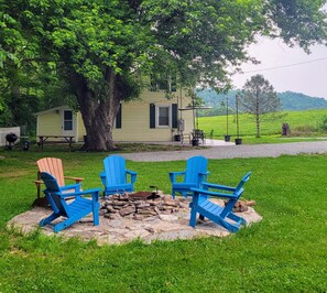 Un espace extérieur rustique aménagé avec des chaises Adirondack disposées autour d’un foyer en pierre, une petite structure en bois et un champ herbeux avec des arbres en arrière-plan.